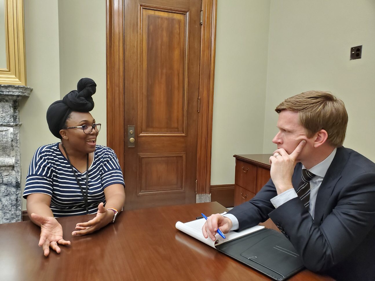 Ally sitting at a table talking to male staffer. Ally’s headwrap looks off brand