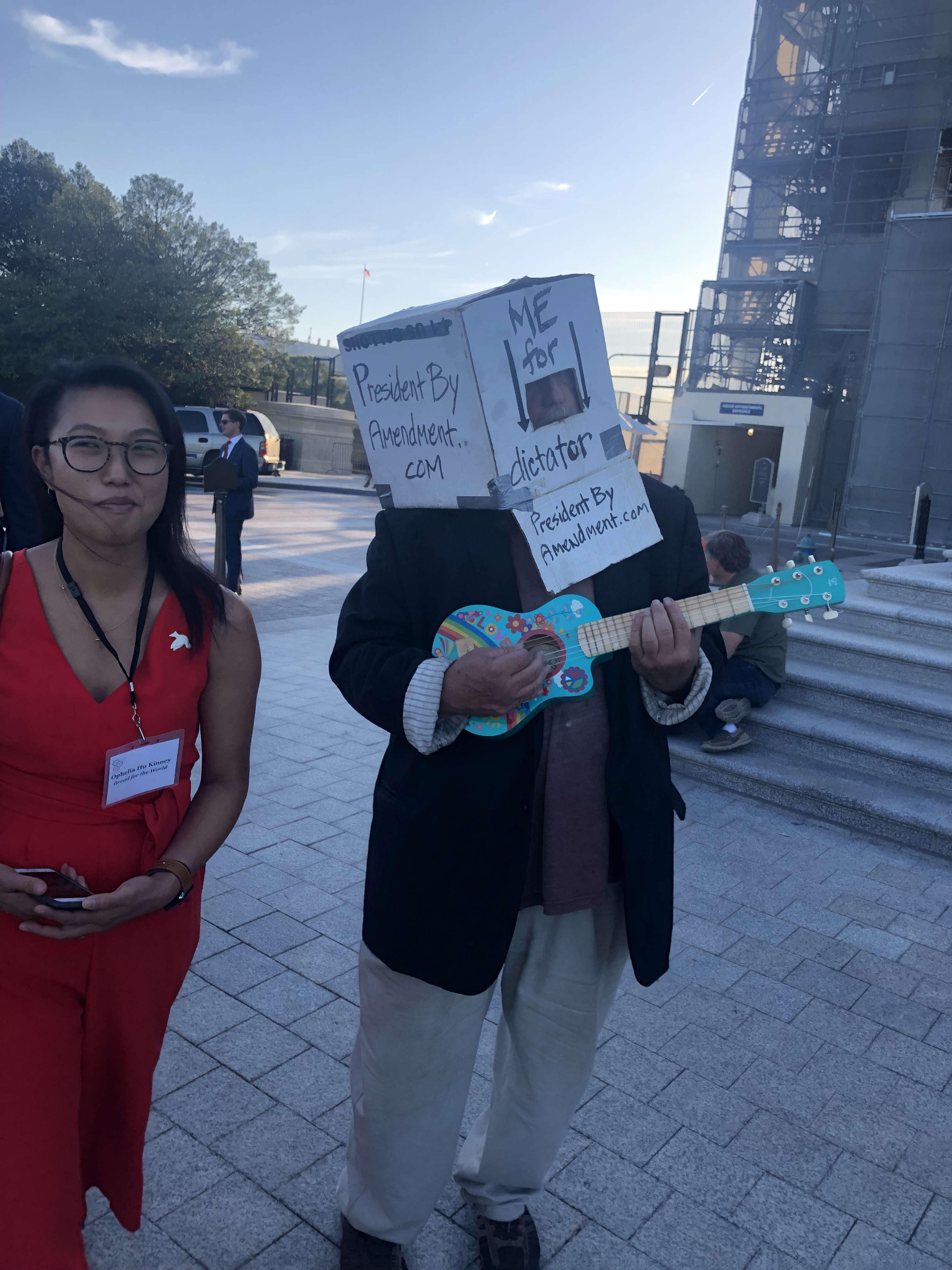 Man with box on his head playing a seafoam green ukulele with a rainbow on it . One side of box says presidentbyammendment.Com. The other says Me for dictator. Asian American woman wearinf red romper walks past attempting not to laugh