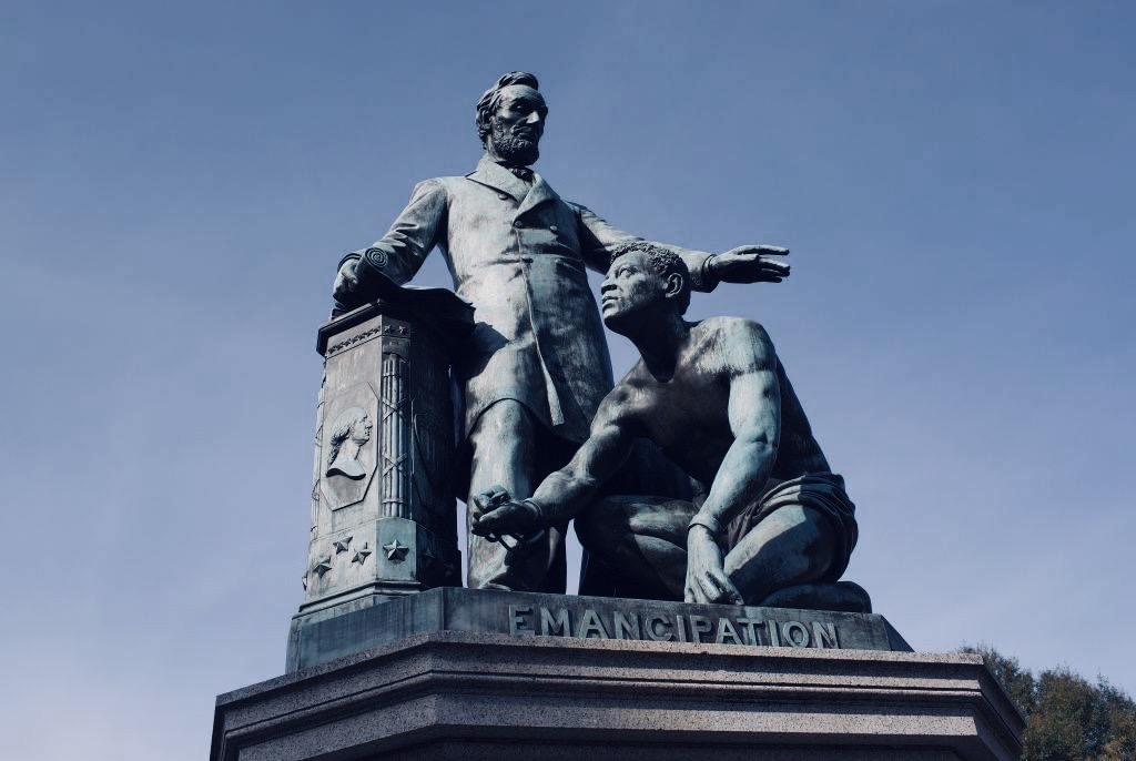 A statue of Abraham Lincoln with his hand on a pillar that has the silhouette of George Washington. Lincoln is standing with his over a kneeling Black man. The word "Emancipation" is inscribed on the statue's base.